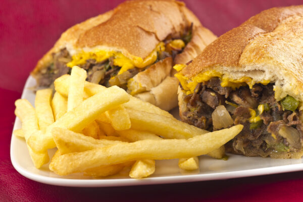 A messy Philly Cheesesteak with onions peppers and mushrooms, fries on the side