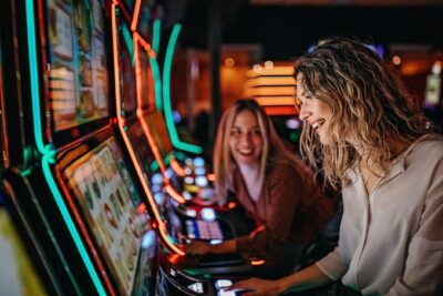 Two women playing slot machines in neon-lit casino.